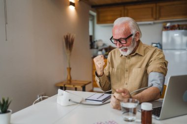 Mutlu, yaşlı, sakallı, sağlıklı beyaz bir erkek, kan basıncı cihazını kullanarak evde tek başına oturup sonuçları ölçüyor. Gerçek insanlar uzay sağlık hizmetleri konseptini kopyalıyor.