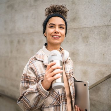 Genç bir kadın yetişkin kız öğrenci ayakta duruyor ya da dışarıda yürüyor dizüstü bilgisayar ve bir fincan kahve tutuyor gerçek bir insan uzayı kopyalıyor mutlu bir gülümseme kulaklıkla gün içinde kendine güveniyor.