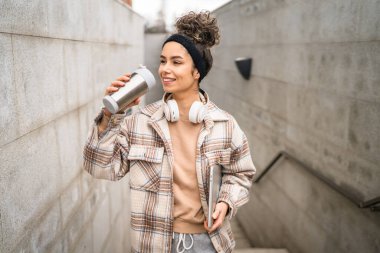 Genç bir kadın yetişkin kız öğrenci ayakta duruyor ya da dışarıda yürüyor dizüstü bilgisayar ve bir fincan kahve tutuyor gerçek bir insan uzayı kopyalıyor mutlu bir gülümseme kulaklıkla gün içinde kendine güveniyor.