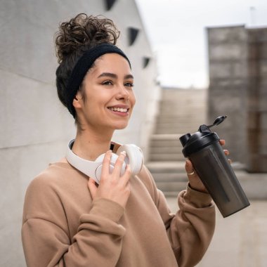 Kulaklıklı bir genç kadın eğitim için hazırlanıyor. Su içmek için destek şişesi tutuyor. Açık havada güzel beyaz kadın nesli.