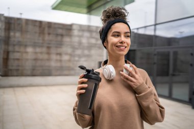 Kulaklıklı bir genç kadın eğitim için hazırlanıyor. Su içmek için destek şişesi tutuyor. Açık havada güzel beyaz kadın nesli.