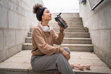 Kulaklıklı bir genç kadın eğitim için hazırlanıyor. Su içmek için destek şişesi tutuyor. Açık havada güzel beyaz kadın nesli.