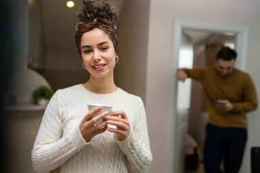 One woman young caucasian female standing in her apartment with cup of coffee while her husband or boyfriend stand in background daily morning evening couple routine real people copy space happy smile