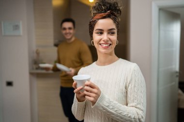 One woman young caucasian female standing in her apartment with cup of coffee while her husband or boyfriend is in the kitchen daily morning evening couple routine happy smile real people copy space