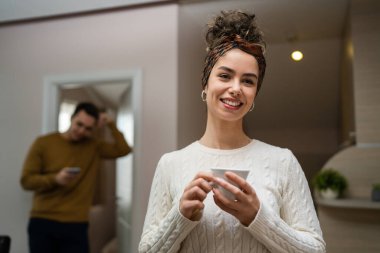 One woman young caucasian female standing in her apartment with cup of coffee while her husband or boyfriend stand in background daily morning evening couple routine real people copy space happy smile