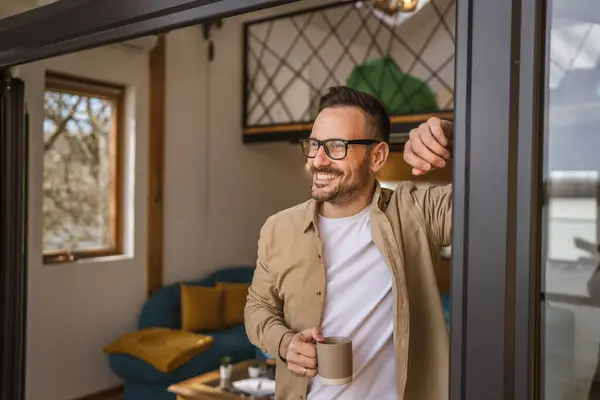 One man adult caucasian male with beard and eyeglasses stand on balcony at door in day happy smile hold cup of coffee copy space morning daily routine concept