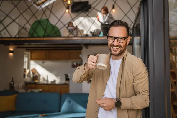 One man adult caucasian male with beard and eyeglasses stand on balcony at door in day happy smile hold cup of coffee copy space morning daily routine concept