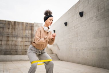 Bir yetişkin beyaz kadın sporcu şehrin dışında esmer ve sağlıklı esmer kadınlar için esneme konseptinde esnemiş beton arka planda elastik lastik direnç bantları ile eğitim görüyor.