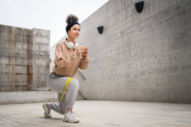 Bir yetişkin beyaz kadın sporcu şehrin dışında esmer ve sağlıklı esmer kadınlar için esneme konseptinde esnemiş beton arka planda elastik lastik direnç bantları ile eğitim görüyor.