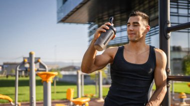 Beyaz tenli genç bir erkek sporcu açık hava spor salonunda çalışırken fren yapıyor. Elinde destek malzemesi tutuyor. Kendine güveni tam, güçlü, güçlü bir kopya alanı var.