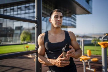 Beyaz tenli genç bir erkek sporcu açık hava spor salonunda çalışırken fren yapıyor. Elinde destek malzemesi tutuyor. Kendine güveni tam, güçlü, güçlü bir kopya alanı var.