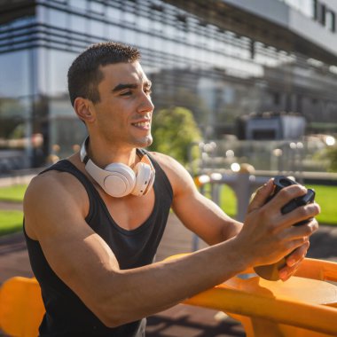Beyaz tenli genç bir erkek sporcu açık hava spor salonunda çalışırken fren yapıyor. Elinde destek malzemesi tutuyor. Kendine güveni tam, güçlü, güçlü bir kopya alanı var.