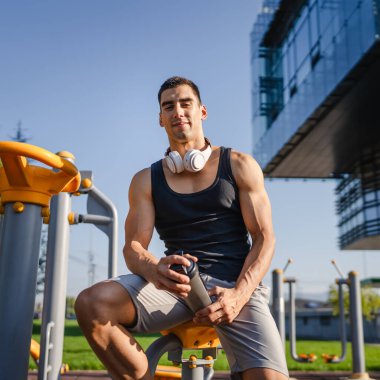 Beyaz tenli genç bir erkek sporcu açık hava spor salonunda çalışırken fren yapıyor. Elinde destek malzemesi tutuyor. Kendine güveni tam, güçlü, güçlü bir kopya alanı var.