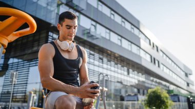 Beyaz tenli genç bir erkek sporcu açık hava spor salonunda çalışırken fren yapıyor. Elinde destek malzemesi tutuyor. Kendine güveni tam, güçlü, güçlü bir kopya alanı var.