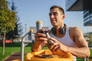 Beyaz tenli genç bir erkek sporcu açık hava spor salonunda çalışırken fren yapıyor. Elinde destek malzemesi tutuyor. Kendine güveni tam, güçlü, güçlü bir kopya alanı var.