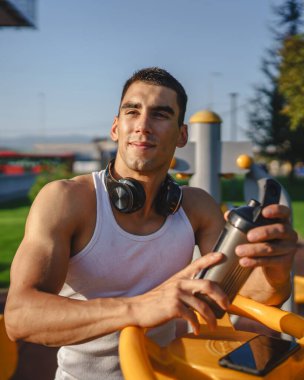 Beyaz tenli genç bir erkek sporcu açık hava spor salonunda çalışırken fren yapıyor. Elinde destek malzemesi tutuyor. Kendine güveni tam, güçlü, güçlü bir kopya alanı var.