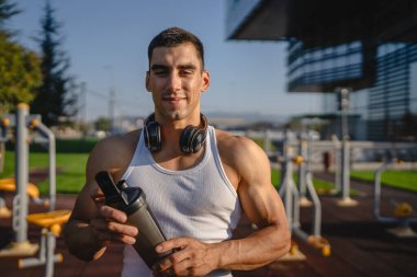 Beyaz tenli genç bir erkek sporcu açık hava spor salonunda çalışırken fren yapıyor. Elinde destek malzemesi tutuyor. Kendine güveni tam, güçlü, güçlü bir kopya alanı var.