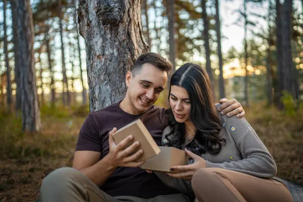 Two people young adult caucasian man and woman couple boyfriend and girlfriend or husband and wife give gift box present while sit in nature forest park celebrate romantic love real people copy space