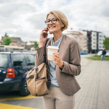 Kafkasyalı, gözlüklü, yetişkin bir kadın otobüs durağında duruyor. Akıllı telefon kullanıyor, telefonla konuşuyor. Gerçek bir insanın işe geç gelmesini bekliyor.