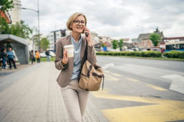 Kafkasyalı, gözlüklü, yetişkin bir kadın otobüs durağında duruyor. Akıllı telefon kullanıyor, telefonla konuşuyor. Gerçek bir insanın işe geç gelmesini bekliyor.