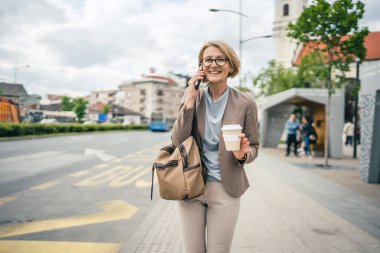 Kafkasyalı, gözlüklü, yetişkin bir kadın otobüs durağında duruyor. Akıllı telefon kullanıyor, telefonla konuşuyor. Gerçek bir insanın işe geç gelmesini bekliyor.