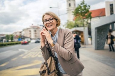 Kafkasyalı, gözlüklü, yetişkin bir kadın otobüs durağında duruyor. Akıllı telefon kullanıyor, telefonla konuşuyor. Gerçek bir insanın işe geç gelmesini bekliyor.