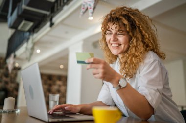 Kıvırcık saçlı, mutlu, yetişkin bir kadın Kafe 'deki laptop bilgisayarından kredi kartı alışverişi yapıyor. Gerçek insanlar e-bankacılık internet indirimi yapıyor.