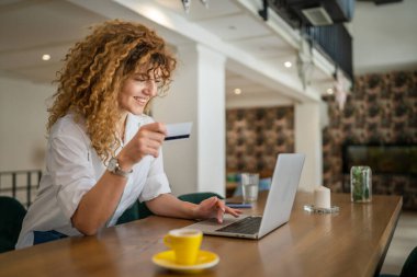 Kıvırcık saçlı, mutlu, yetişkin bir kadın Kafe 'deki laptop bilgisayarından kredi kartı alışverişi yapıyor. Gerçek insanlar e-bankacılık internet indirimi yapıyor.