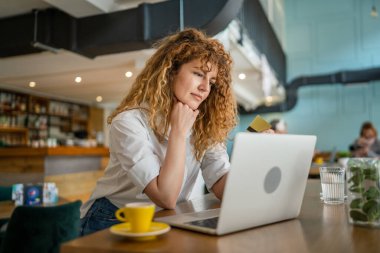 Kıvırcık saçlı, mutlu, yetişkin bir kadın Kafe 'deki laptop bilgisayarından kredi kartı alışverişi yapıyor. Gerçek insanlar e-bankacılık internet indirimi yapıyor.