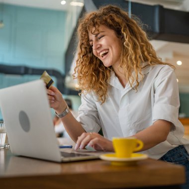 Kıvırcık saçlı, mutlu, yetişkin bir kadın Kafe 'deki laptop bilgisayarından kredi kartı alışverişi yapıyor. Gerçek insanlar e-bankacılık internet indirimi yapıyor.