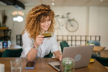 Kıvırcık saçlı, mutlu, yetişkin bir kadın Kafe 'deki laptop bilgisayarından kredi kartı alışverişi yapıyor. Gerçek insanlar e-bankacılık internet indirimi yapıyor.