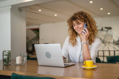 Yetişkin bir kadın Kafkasyalı kadın laptopun başında oturuyor. Telefonla konuşuyor. Mutlu bir gülümseme.