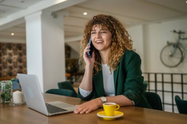 Yetişkin bir kadın Kafkasyalı kadın laptopun başında oturuyor. Telefonla konuşuyor. Mutlu bir gülümseme.