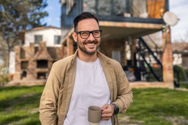 40 yaşında yetişkin bir adam elinde bir fincan çay ya da kahveyle modern evinin önünde güvenle duruyor hayatından memnun ve mutlu görünüyor ve evinin fotokopi alanından gurur duyuyor.