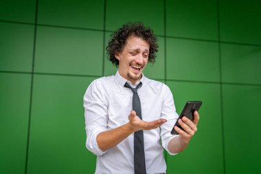 Kafkasyalı bir işadamı dışarıda duruyor mutlu bir gülücük atıyor video oyunları oynuyor, eğleniyor cep telefonu kullanıyor akıllı telefon kullanıyor ve internetteki yetişkin erkeklerin fotokopisini çekiyor.