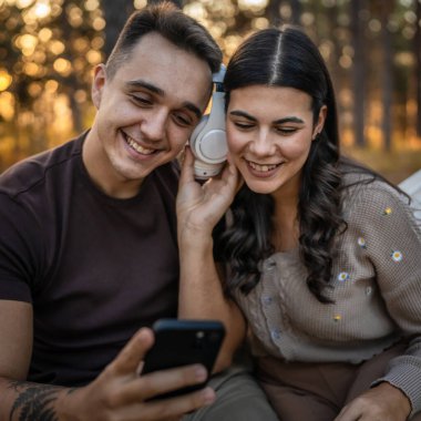 Doğadaki genç erkek ve kadın yetişkin çift kulaklıkla müzik dinliyor cep telefonu kullanıyor ya da video arama uygulaması yapıyor.