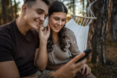 Doğadaki genç erkek ve kadın yetişkin çift kulaklıkla müzik dinliyor cep telefonu kullanıyor ya da video arama uygulaması yapıyor.
