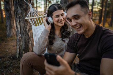 Doğadaki genç erkek ve kadın yetişkin çift kulaklıkla müzik dinliyor cep telefonu kullanıyor ya da video arama uygulaması yapıyor.