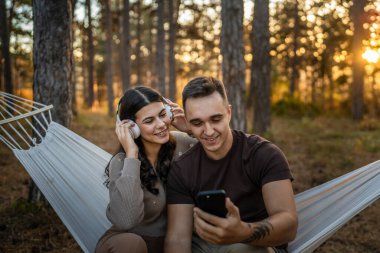 Doğadaki genç erkek ve kadın yetişkin çift kulaklıkla müzik dinliyor cep telefonu kullanıyor ya da video arama uygulaması yapıyor.