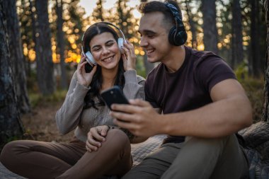Doğadaki genç erkek ve kadın yetişkin çift kulaklıkla müzik dinliyor cep telefonu kullanıyor ya da video arama uygulaması yapıyor.