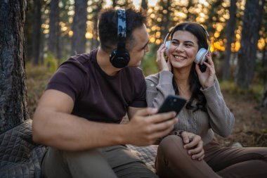 Doğadaki genç erkek ve kadın yetişkin çift kulaklıkla müzik dinliyor cep telefonu kullanıyor ya da video arama uygulaması yapıyor.