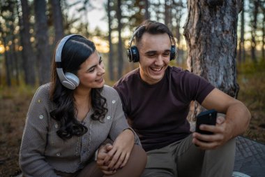 Doğadaki genç erkek ve kadın yetişkin çift kulaklıkla müzik dinliyor cep telefonu kullanıyor ya da video arama uygulaması yapıyor.