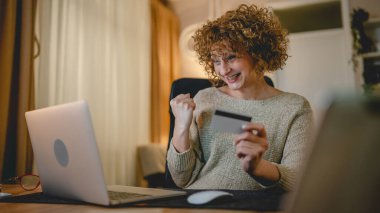 Modern beyaz bir kadın internetten alışveriş yapıyor. Dizüstü bilgisayar ve kredi kartı kullanıyor. Evinden mutlu bir gülümseme.