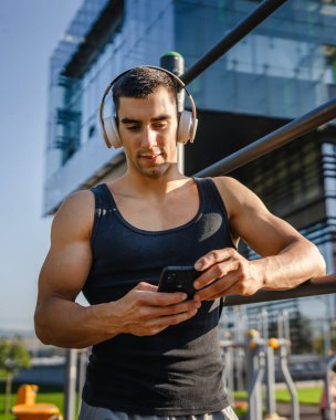Beyaz tenli genç bir erkek açık hava eğitim parkı spor salonunda duruyor. Kulaklıklı cep telefonu kullanıyor. Gerçek insanları eğitmek için internetten mesajlar yolluyor.