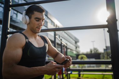 Beyaz tenli genç bir erkek açık hava eğitim parkı spor salonunda duruyor. Kulaklıklı cep telefonu kullanıyor. Gerçek insanları eğitmek için internetten mesajlar yolluyor.