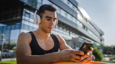 Beyaz tenli genç bir erkek açık hava eğitim parkı spor salonunda duruyor. Kulaklıklı cep telefonu kullanıyor. Gerçek insanları eğitmek için internetten mesajlar yolluyor.