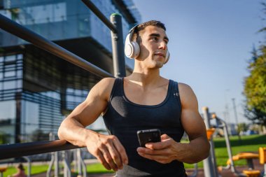 Beyaz tenli genç bir erkek açık hava eğitim parkı spor salonunda duruyor. Kulaklıklı cep telefonu kullanıyor. Gerçek insanları eğitmek için internetten mesajlar yolluyor.