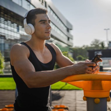 Beyaz tenli genç bir erkek açık hava eğitim parkı spor salonunda duruyor. Kulaklıklı cep telefonu kullanıyor. Gerçek insanları eğitmek için internetten mesajlar yolluyor.