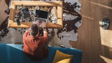 Üst görüşlü adam ev işlerinde kanepeye oturup bilgisayarın kumandasını kullanıyor.