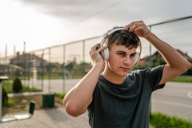 Beyaz tenli genç bir erkek açık hava eğitim parkı spor salonunda duruyor. Kulaklıklı cep telefonu kullanıyor. Gerçek insanları eğitmek için internetten mesajlar yolluyor.